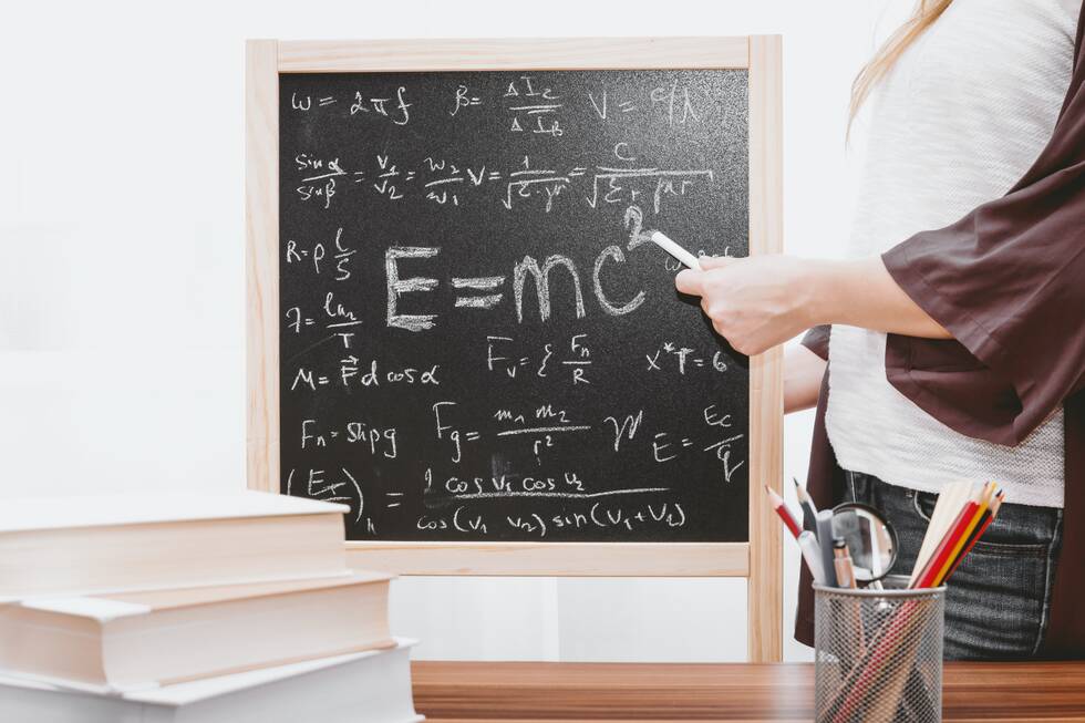 Female hand holding white cahlk and pointing to a small blackboard with calculations and E=MC2 equation on it