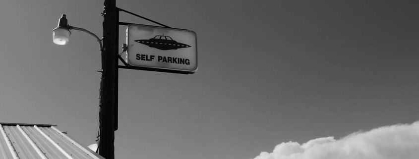 B&W photo of a lamp-post with a 'self-parking' sign that also has a drawing of a spaceship on it.